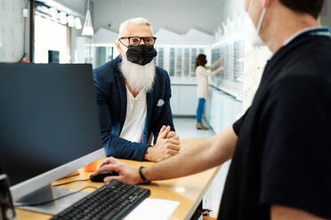 Elderly male in mask standing at desk and talking to optician in optical store - ADSF24274