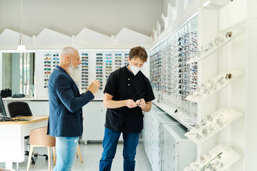 Side view of professional optician in mask helping senior male choosing glasses while working in optical store - ADSF24276