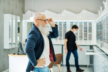 Seitenansicht eines professionellen Optikers mit Maske, der einem älteren Mann bei der Auswahl einer Brille hilft, während er in einem optischen Geschäft arbeitet - ADSF24277