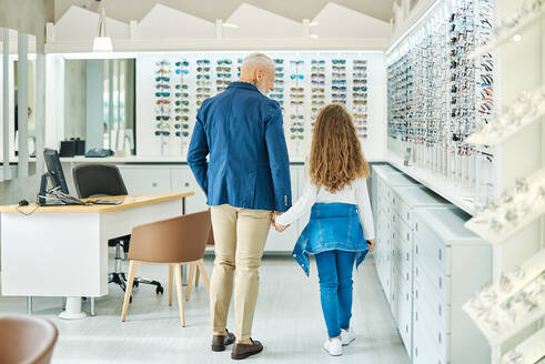 Rückenansicht eines stilvollen älteren Mannes, der die Hand seiner Enkelin im Teenageralter hält, während er in einem Optikgeschäft steht - ADSF24280