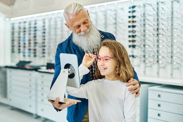 Fröhlicher älterer Mann, der einen Spiegel hält, während ein junges Mädchen in einem modernen Optikgeschäft eine Brille anprobiert - ADSF24281