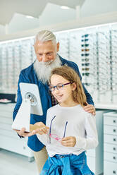 Cheerful senior man holding mirror while teenage girl trying on glasses in modern optical store - ADSF24282