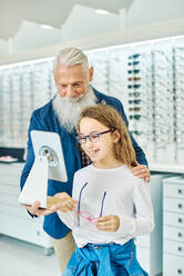 Fröhlicher älterer Mann, der einen Spiegel hält, während ein junges Mädchen in einem modernen Optikgeschäft eine Brille anprobiert - ADSF24282