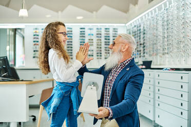 Seitenansicht eines fröhlichen Großvaters und einer Enkelin mit Brille, die sich in einem Optikgeschäft gegenseitig die Daumen drücken - ADSF24283