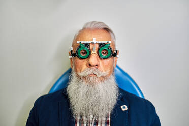 Bearded elderly male in eye test glasses sitting in modern clinic before vision examination and looking at camera - ADSF24285