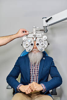 Unkenntlich gemachter Optiker mit Phoropter zur Überprüfung der Sehkraft eines älteren männlichen Patienten in der Klinik - ADSF24286