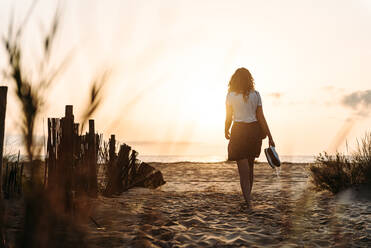 Rückenansicht einer nicht erkennbaren Frau in Sommerkleidung, die am Sandstrand entlang in Richtung Meer läuft, vor dem Hintergrund des Himmels bei Sonnenuntergang - ADSF24289