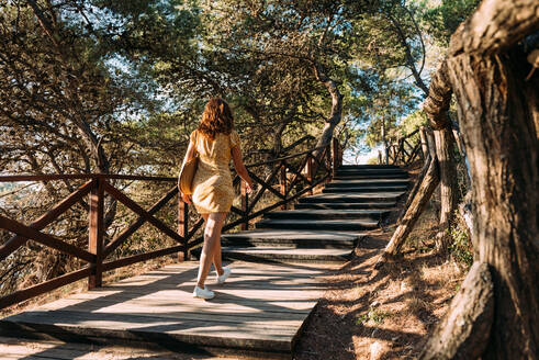 Rückenansicht einer nicht erkennbaren Frau in einem Kleid, die einen Holzweg in der Natur an einem sonnigen Sommertag hinaufgeht - ADSF24291