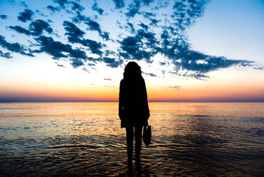 Rückansicht der Silhouette einer nicht erkennbaren Frau, die im Wasser des Meeres steht und den spektakulären Blick auf den farbenfrohen Himmel bei Sonnenuntergang im Sommer bewundert - ADSF24292