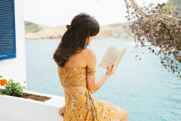 Seitenansicht einer romantischen jungen Frau in Sommerkleidung, die auf einer Terrasse in der Nähe eines Sees sitzt und einen interessanten Roman in einem Buch liest, während sie die Sommerzeit auf dem Land verbringt - ADSF24294