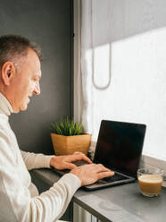 Seitenansicht eines lächelnden Mannes mittleren Alters, der mit einem Netbook und einer Tasse Kaffee in der Küche am Morgen arbeitet - ADSF24314