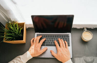 Von oben abgeschnittene unerkennbare männliche Person mittleren Alters, die mit einem Netbook und einer Tasse Kaffee in der Küche am Morgen am Tresen arbeitet - ADSF24315