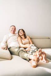 Delighted middle aged couple sitting on comfortable sofa in living room at home while smiling and enjoying weekend together - ADSF24321