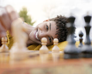 Von unten optimistische afroamerikanische Frau, die an einem Tisch im Park sitzt und Schach spielt, während sie in die Kamera schaut - ADSF24332