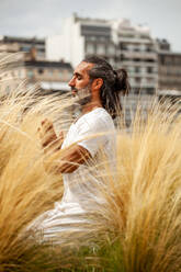 Unrasierter hispanischer Mann in weißer Kleidung sitzt mit geschlossenen Augen während der Meditation im goldenen Gras bei Tageslicht - ADSF24344