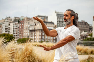 Mature unshaven ethnic male in white outfit looking forward while practicing yoga against city buildings - ADSF24347