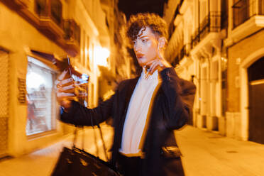Junge trendige Transgender-Mann mit Make-up berühren Wange während der Aufnahme von Selbstporträt auf Handy in der Stadt in der Nacht - ADSF24358