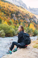Seitenansicht einer Wanderin mit Rucksack, die auf einem Felsen in der Nähe eines Flusses in den Pyrenäen sitzt, während sie ihren Urlaub im Nationalpark Ordesa y Monte Perdido genießt - ADSF24363