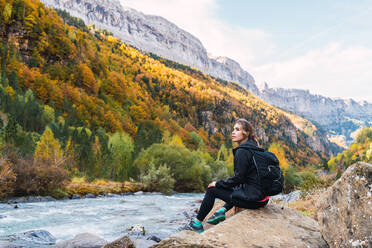 Seitenansicht einer Wanderin mit Rucksack, die auf einem Felsen in der Nähe eines Flusses in den Pyrenäen sitzt, während sie ihren Urlaub im Nationalpark Ordesa y Monte Perdido genießt - ADSF24364