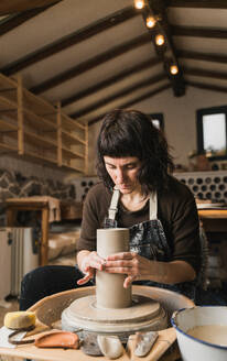 Fokussierte Kunsthandwerkerin, die auf der Töpferscheibe Geschirr aus Ton herstellt, während sie im Atelier arbeitet - ADSF24369