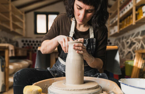 Focused female artisan creating clay tableware on pottery wheel while working in art studio - ADSF24371