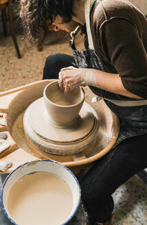 Side view of focused unrecognizable female artisan using pottery wheel and creating handmade earthenware in workshop - ADSF24372