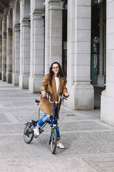 Fröhliche Frau im Mantel, die auf einem Fahrrad gegen ein altes Gebäude mit Säulen in der Stadt wegschaut - ADSF24375