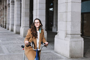 Cheerful woman in coat looking away on bike against old building with columns in town - ADSF24376