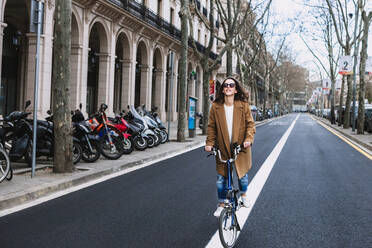 Cheerful woman in coat looking away on bike against old building with columns in town - ADSF24378