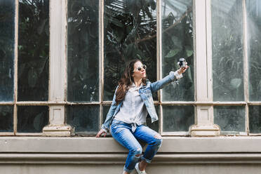 Coole Frau mit Sonnenbrille und Jeanskleidung, die ein Selbstporträt mit einer Fotokamera aufnimmt, während sie vor dem Fenster einer Orangerie in der Stadt sitzt - ADSF24379