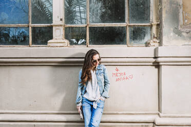 Junge Frau in trendigem Jeans-Outfit und Sonnenbrille steht mit der Hand in der Tasche in der Nähe eines schäbigen Gebäudes in der Stadt - ADSF24380