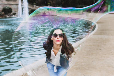 Unbeschwerte Frau in modischer Kleidung und Sonnenbrille steht in der Nähe eines Brunnens im Park und bläst Seifenblasen - ADSF24382