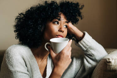 Pensive African American female with mug of beverage sitting on sofa at home and looking away in contemplation - ADSF24387