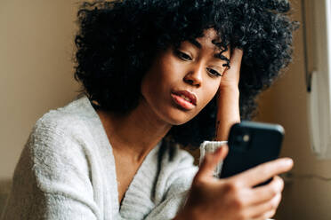 Side view of thoughtful African American female sitting on soft couch in living room and browsing mobile phone at weekend at home - ADSF24388