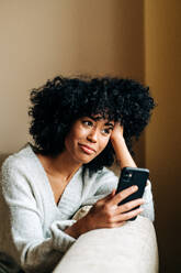 Seitenansicht einer glücklichen afroamerikanischen Frau, die auf einer weichen Couch im Wohnzimmer sitzt und am Wochenende zu Hause auf ihrem Mobiltelefon surft - ADSF24390