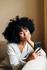 Seitenansicht einer nachdenklichen afroamerikanischen Frau, die auf einer weichen Couch im Wohnzimmer sitzt und am Wochenende zu Hause auf ihrem Mobiltelefon surft - ADSF24391