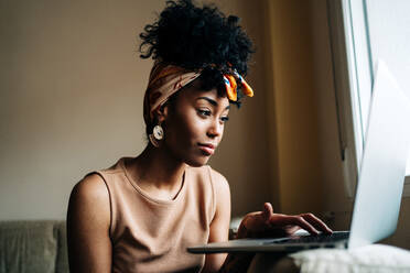 African American female in stylish outfit sitting on sofa at home and typing on netbook while working on remote project - ADSF24402