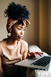 Afroamerikanische Frau in stilvollem Outfit sitzt zu Hause auf dem Sofa und tippt auf einem Netbook, während sie an einem Fernprojekt arbeitet - ADSF24403