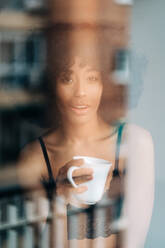 Blick durch das Fenster auf eine afroamerikanische Frau in einem Spitzentop und mit einer Tasse Getränk, die in die Kamera schaut - ADSF24415