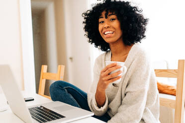 Zufriedene afroamerikanische Freiberuflerin mit einem Becher Heißgetränk, die mit ihrem Laptop am Tisch sitzt und in die Kamera schaut, während sie im Heimbüro arbeitet - ADSF24418