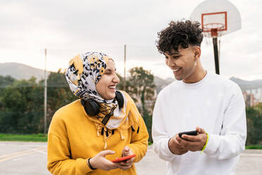 Vergnügte Freunde, die auf einem Basketballplatz stehen und auf ihren Smartphones surfen, während sie lachen und Spaß in der Stadt haben - ADSF24468
