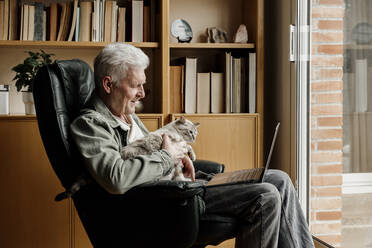 Senior man with laptop holding cat while sitting on chair at home - AFVF08747