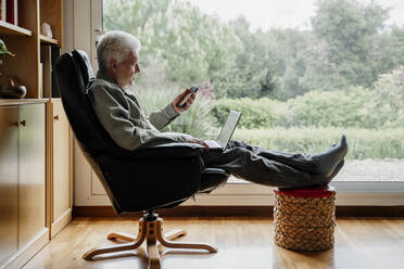 Senior man using mobile phone while sitting on chair at home - AFVF08752