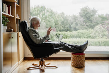 Senior man waving on video call through laptop at home - AFVF08753