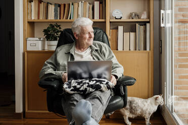 Lächelnder älterer Mann mit Katze, der durch das Fenster seines Hauses schaut - AFVF08757