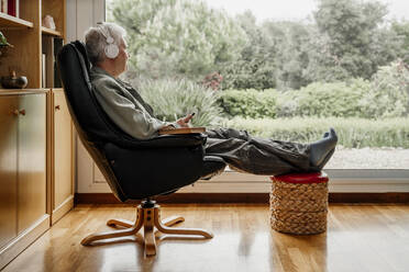 Senior man looking through window while listening music at home - AFVF08761