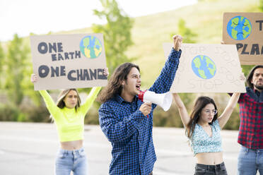 Männlicher Aktivist mit Megaphon, der eine Rede zum Klimawandel hält, mit Demonstranten im Hintergrund auf einem Fußweg - JCCMF02432