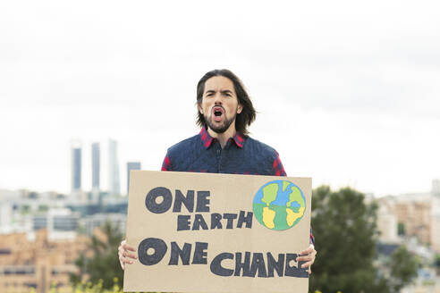 Langhaariger Mann mit Plakat, der vor dem Himmel über den Klimawandel schreit - JCCMF02454