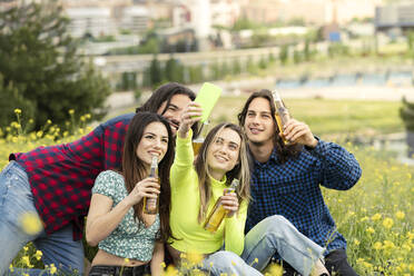 Woman taking selfie with male and female friends while having beer on field in nature - JCCMF02459