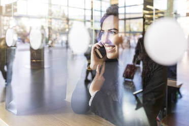 Lächelnde Geschäftsfrau, die wegschaut, während sie am Tisch mit ihrem Smartphone telefoniert - WPEF04456